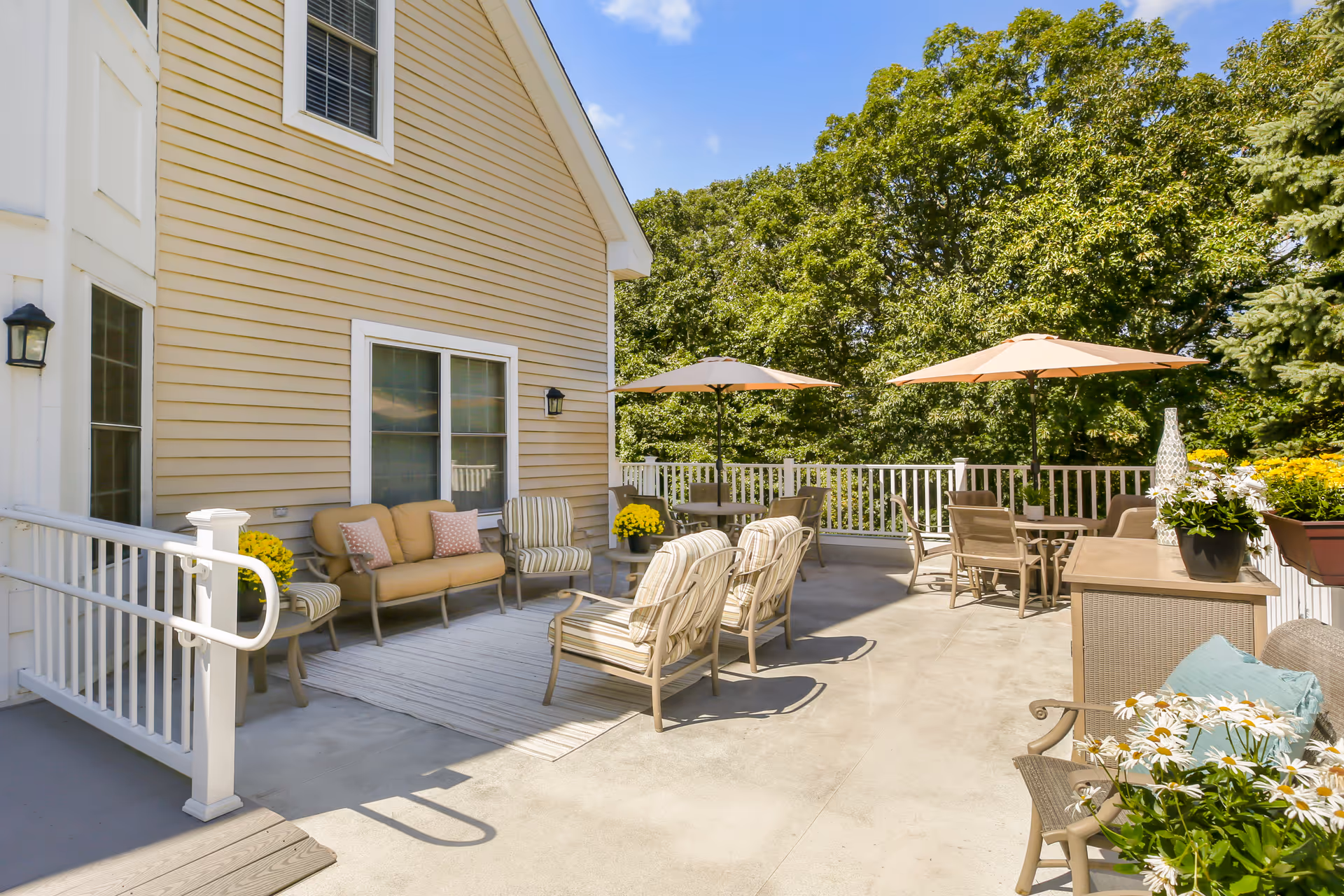 Outdoor patio area at Brookdale Attleboro with beige siding building wall, cushioned chairs, sofas with pillows, tables with umbrellas, potted flowers, and surrounded by green trees under a clear blue sky.