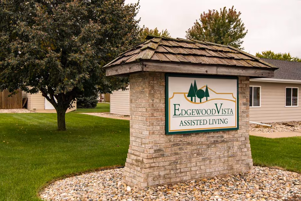 A brick entry sign with a shingled roof that reads "Edgewood Vista Assisted Living" in front of a lawn, trees, and a single-story building.
