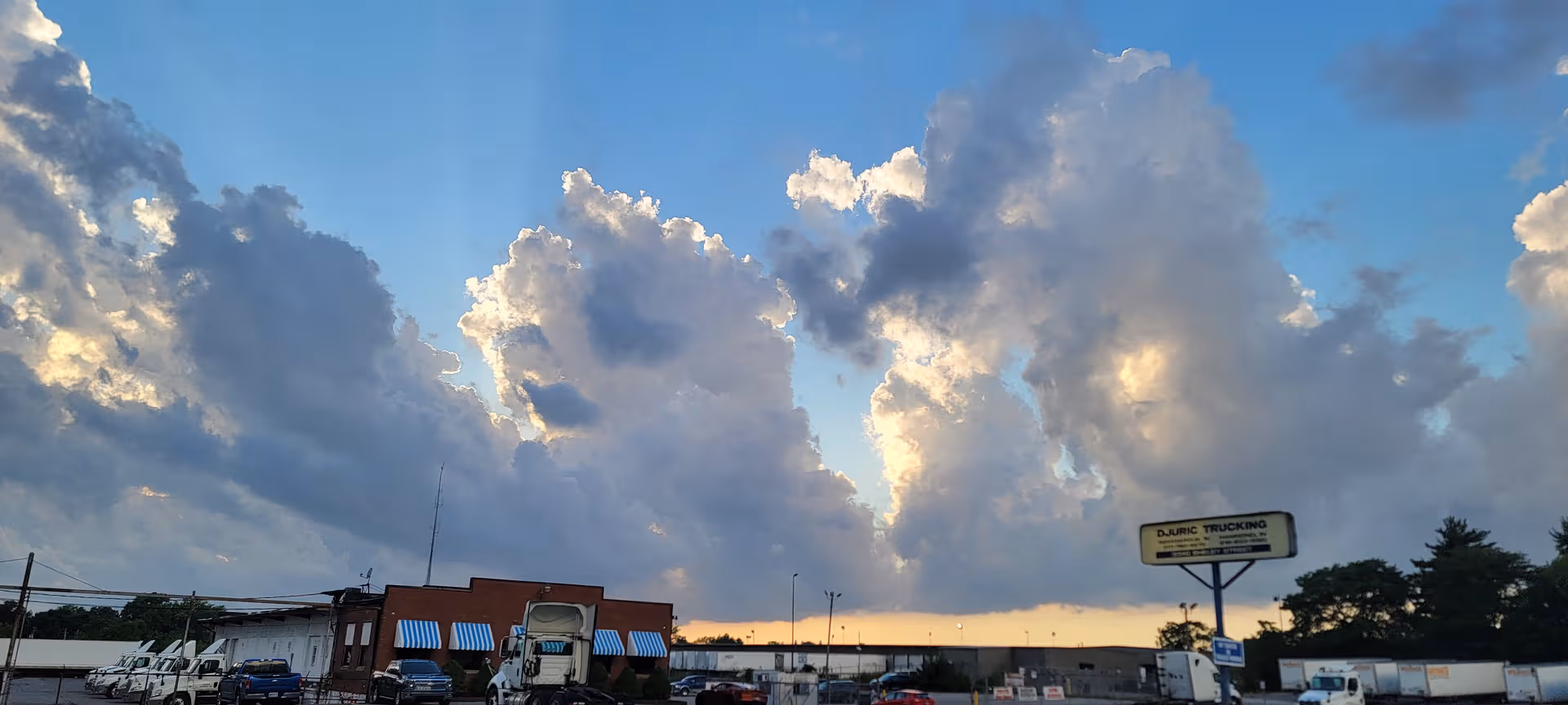 Wide exterior view of a row of storefronts, trucks, and a sunset sky.