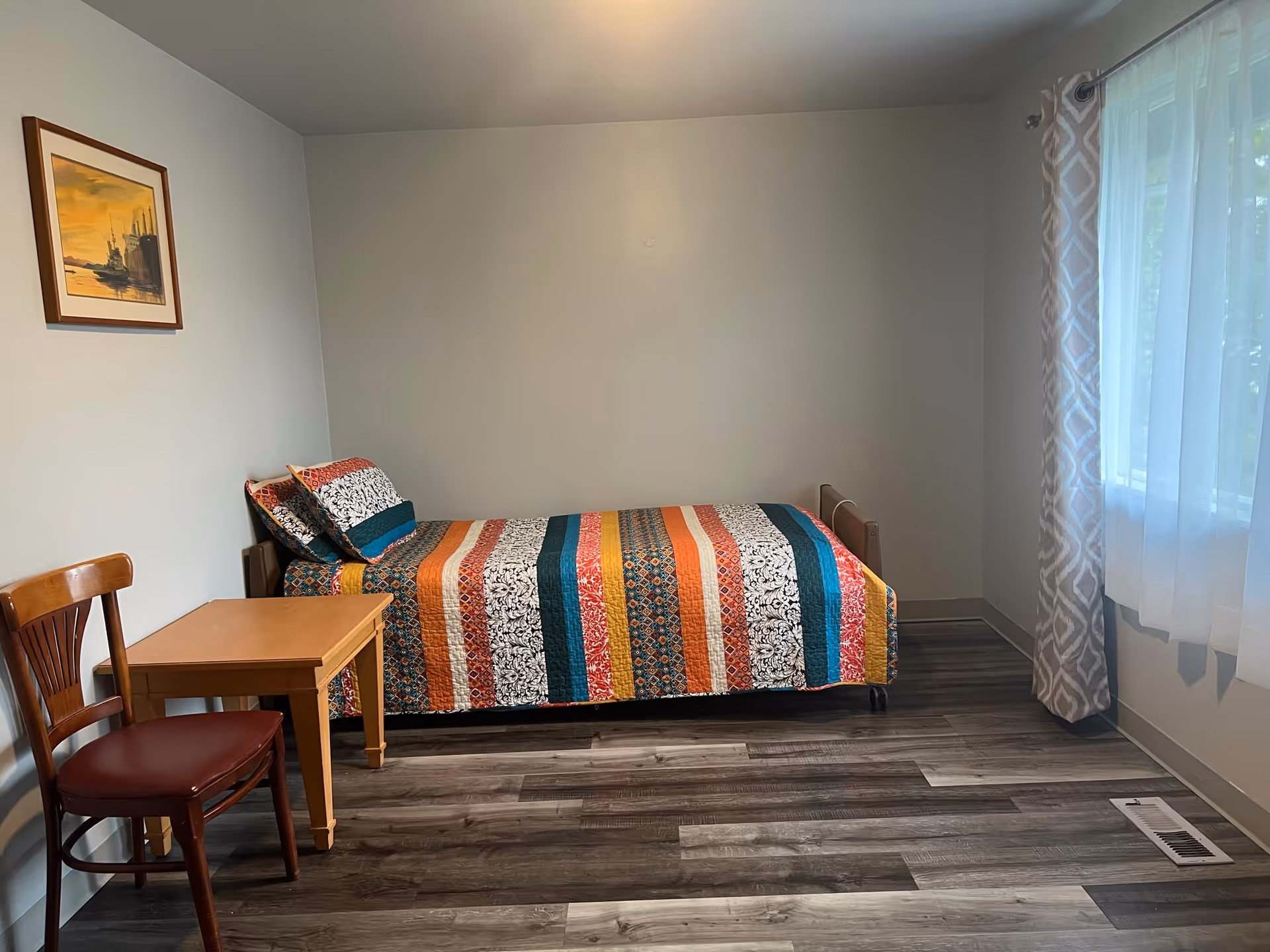 Single bedroom with colorful quilted bed, table, chair, and window with sheer curtains.