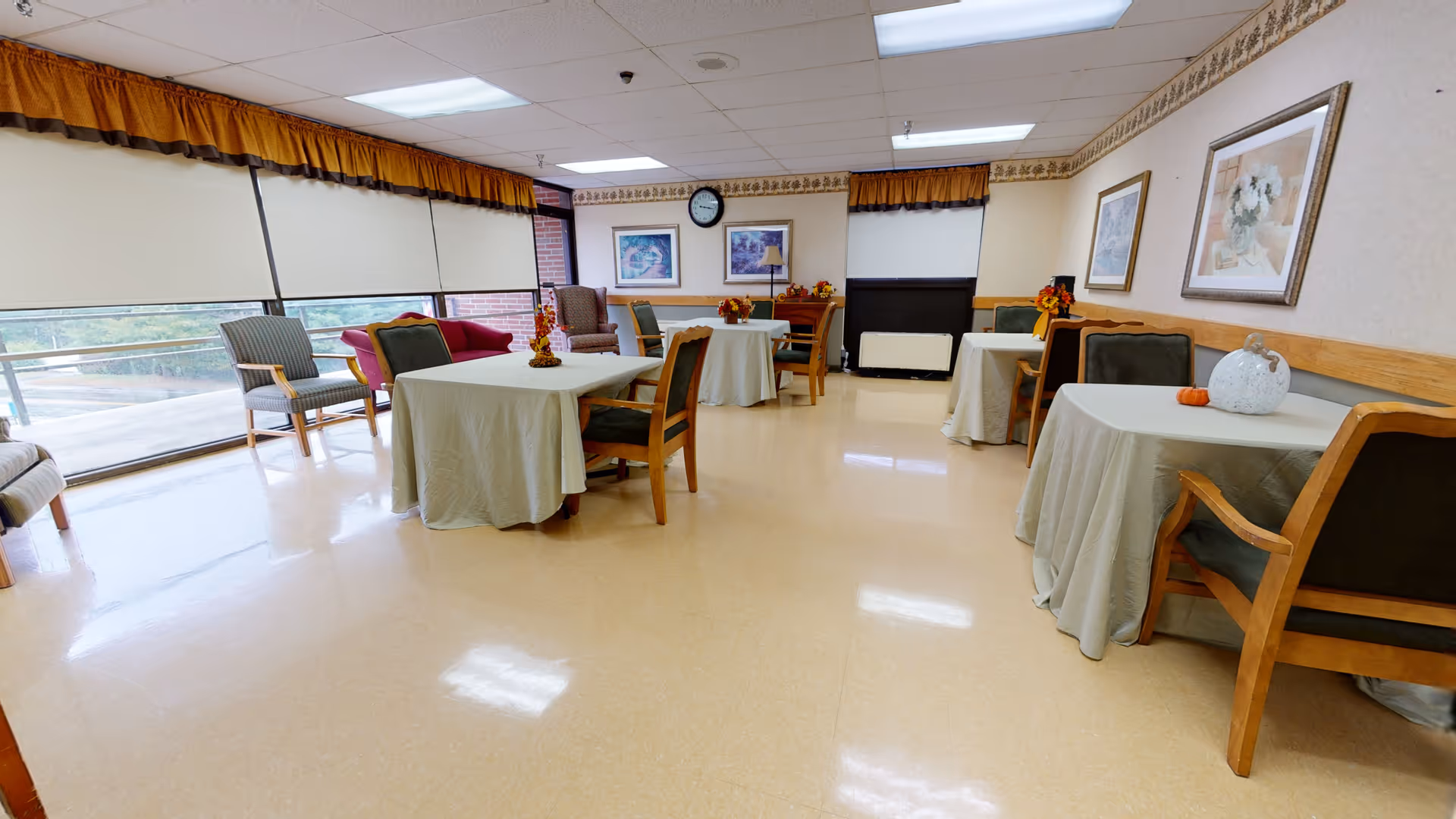 A bright and clean dining room with several tables covered in light-colored tablecloths, each surrounded by wooden chairs with green cushions. The room has large windows with white blinds and orange valances, framed artwork on the walls, and a clock above a small cabinet. The floor is a polished light beige color, and there are decorative items on the tables including small pumpkins and floral arrangements.