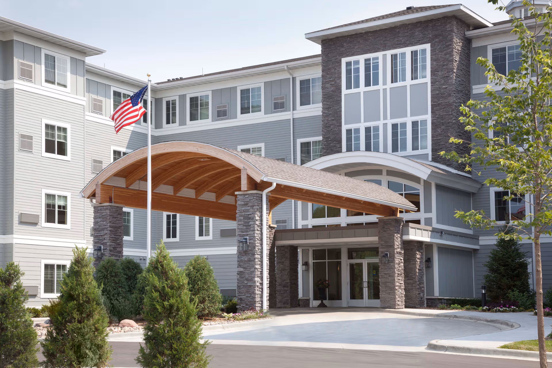 Modern senior living facility exterior with a curved wooden canopy, stone columns, and American flag.