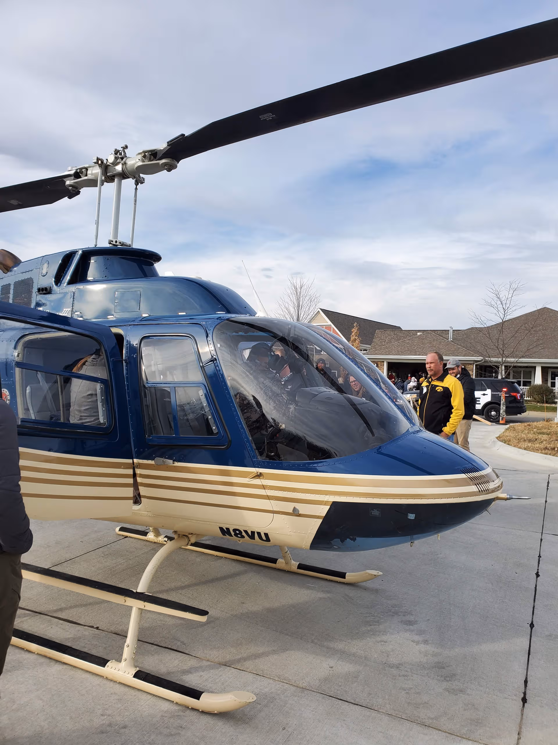 Blue helicopter on a driveway with people gathered nearby at a facility.