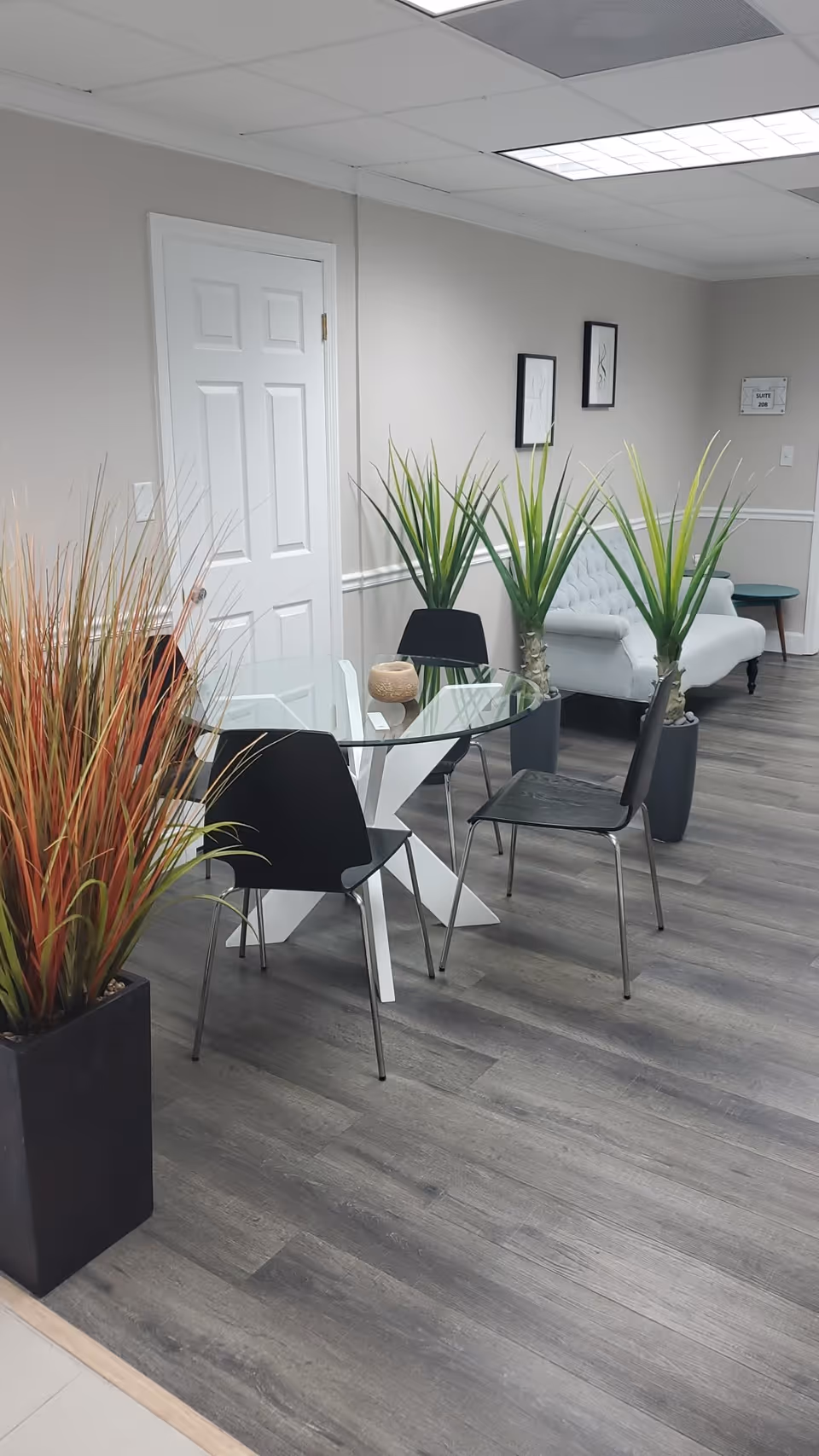 Interior lounge area with glass round table, black chairs, and tall potted plants.