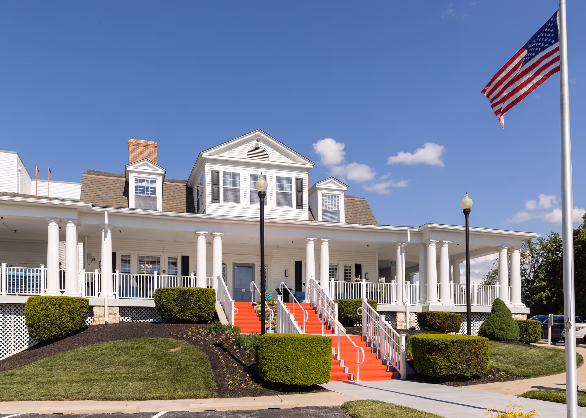 Front exterior view of The Terraces at Westminster building with white columns, a red carpeted staircase, neatly trimmed bushes, an American flag on a flagpole, and a clear blue sky with a few clouds.