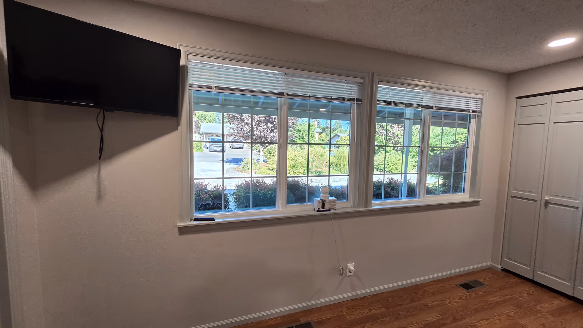 Empty bedroom with a wall-mounted TV, large window, blinds, and white closet doors.