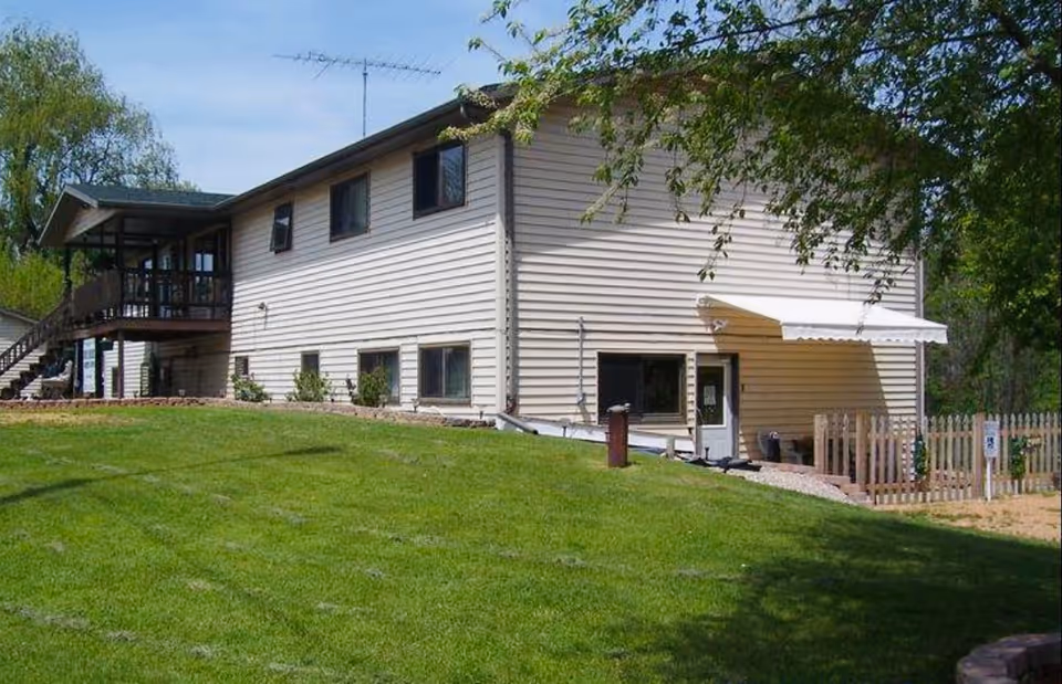 Exterior view of a two-story beige building with multiple windows and a small fenced area with a white awning over a door. The building is surrounded by a well-maintained green lawn and some trees.
