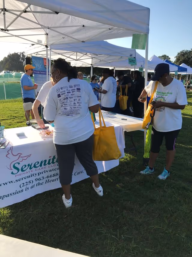 Outdoor community fair with white tents, volunteers, and Serenity Private Care promotional table.