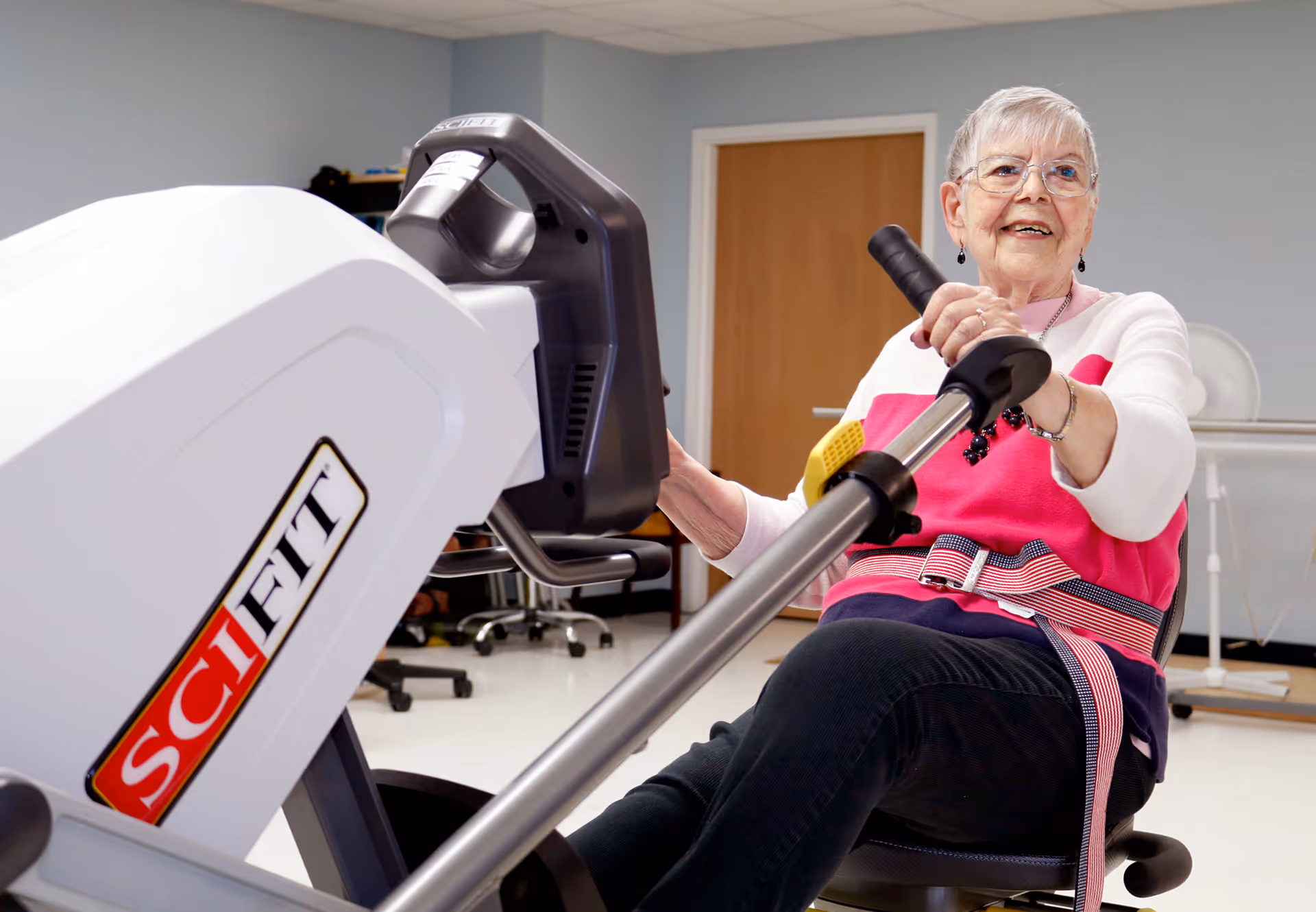 An elderly woman with short gray hair and glasses is exercising on a SciFit rowing machine in a room with light blue walls and a wooden door. She is wearing a pink and white sweater, black pants, and a safety belt, and she is smiling while holding the rowing machine handles.