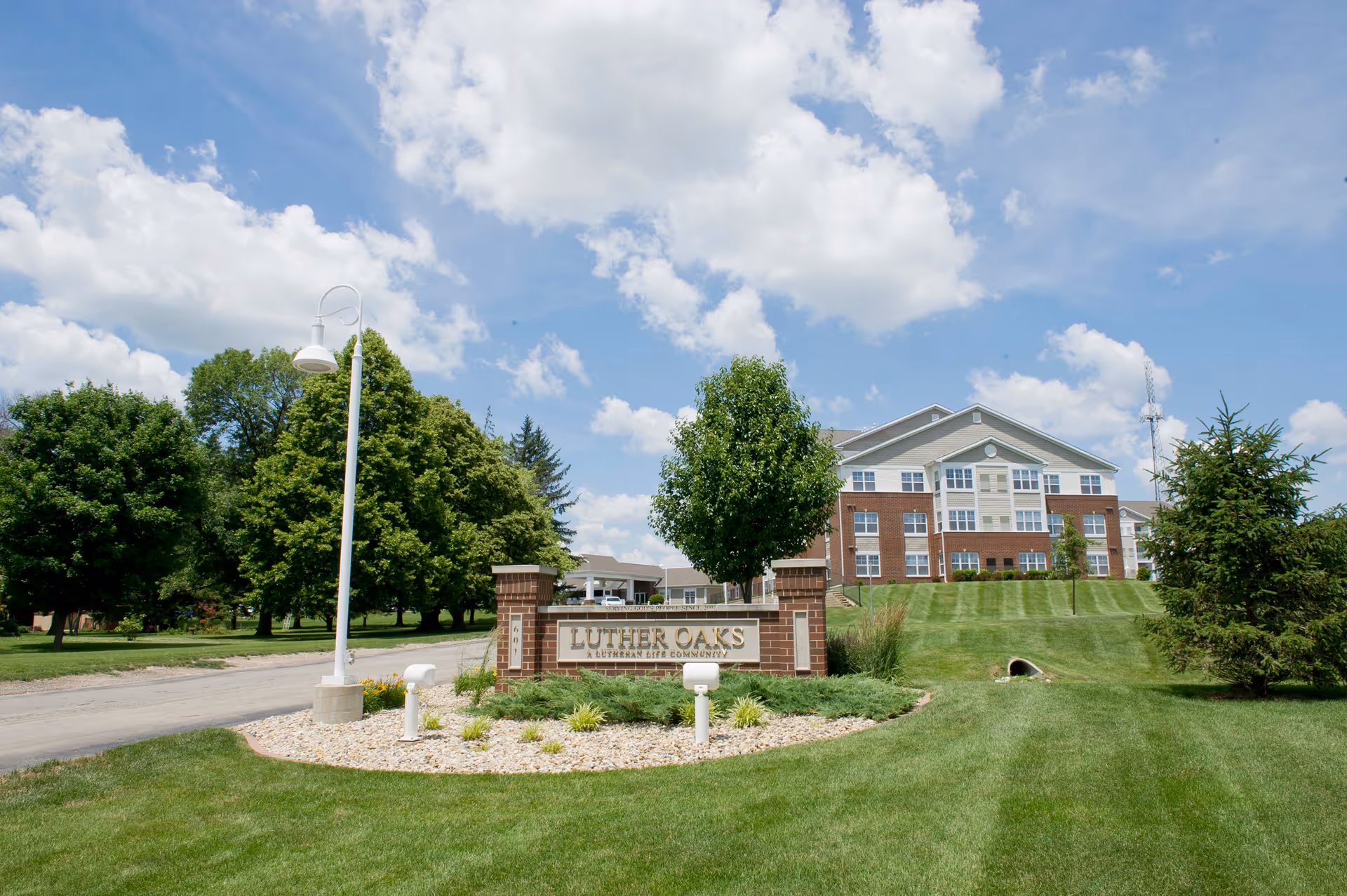 Entrance sign for Luther Oaks on a landscaped lawn with a multi-story senior living building and trees under a blue sky.