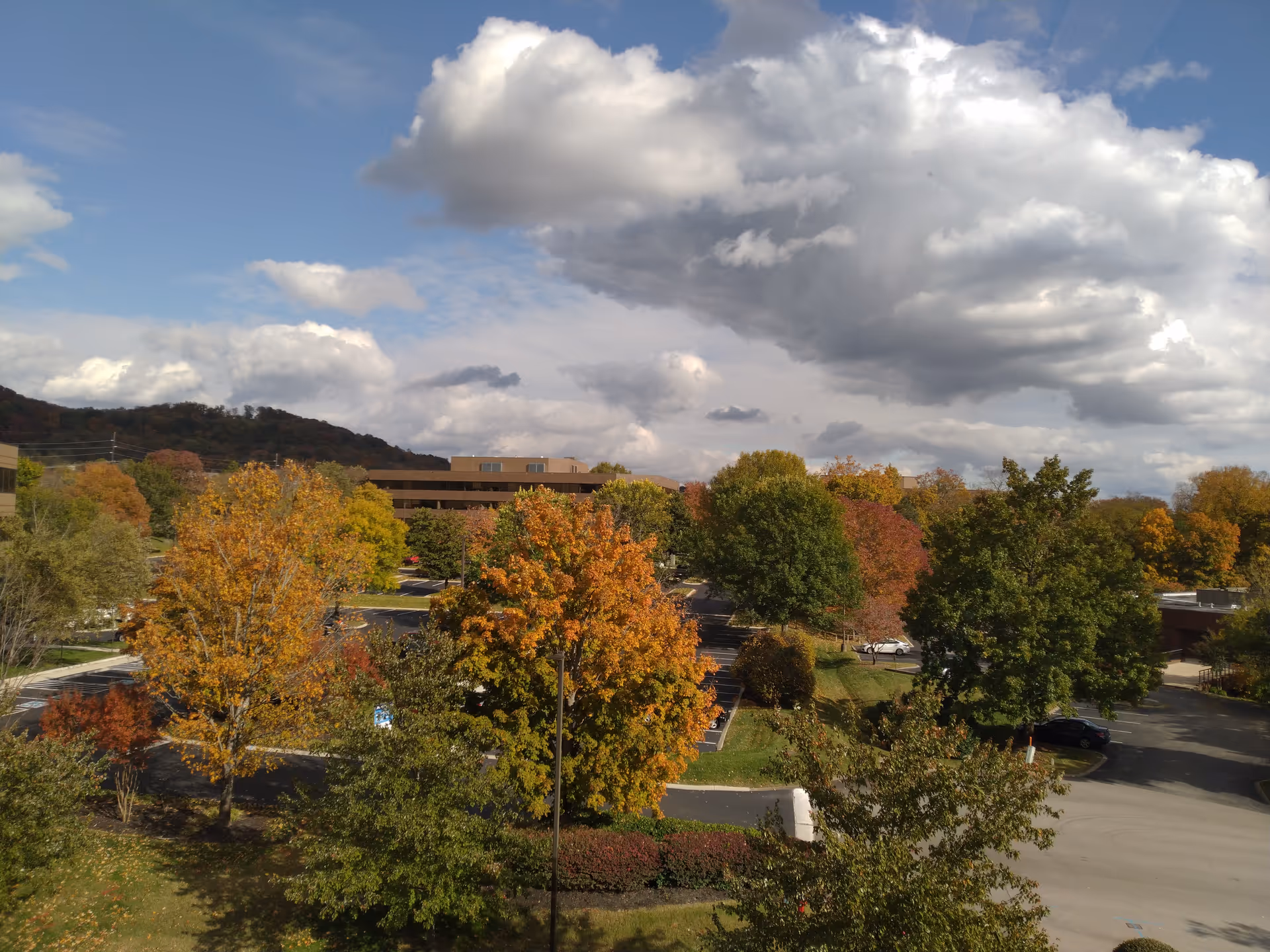 Autumn trees surround a parking lot and a low-rise building under a cloudy sky.