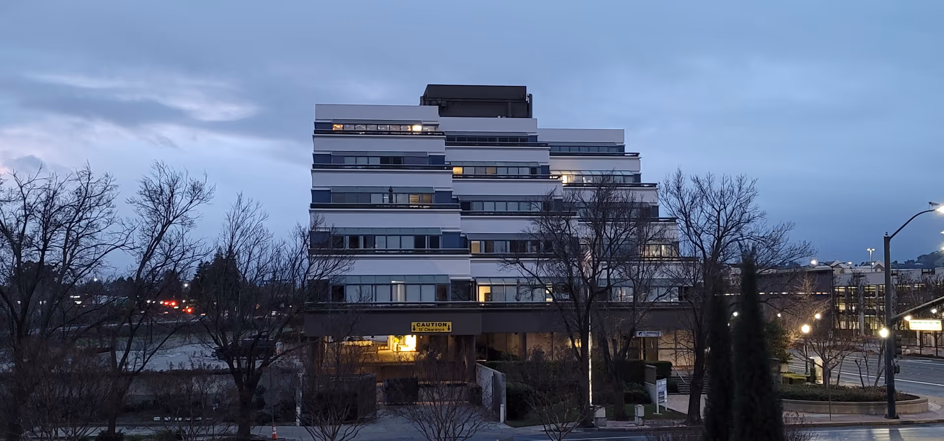 Modern multi-story building with stacked balconies at dusk, surrounded by leafless trees.