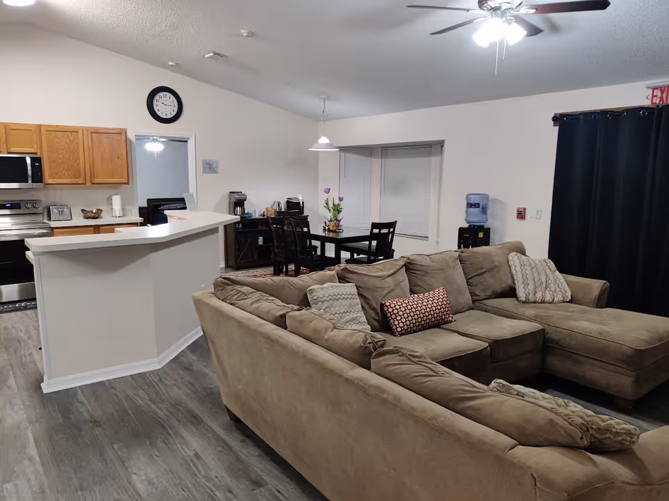 Interior view of a living area in an assisted living facility showing a large beige sectional sofa with multiple cushions, a dining table with chairs and a flower vase, a kitchen area with wooden cabinets and stainless steel appliances, a wall clock, and a water dispenser near a window with closed blinds.