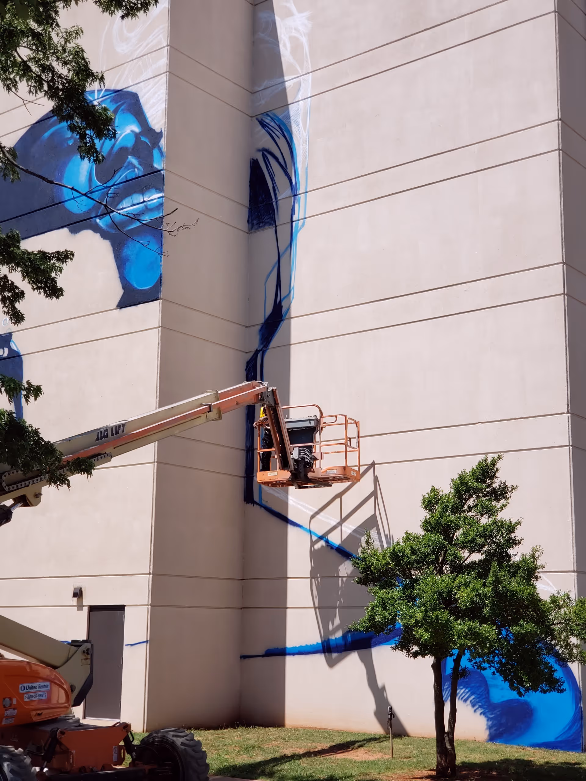 Tall beige building wall with blue mural being painted by an elevated lift.