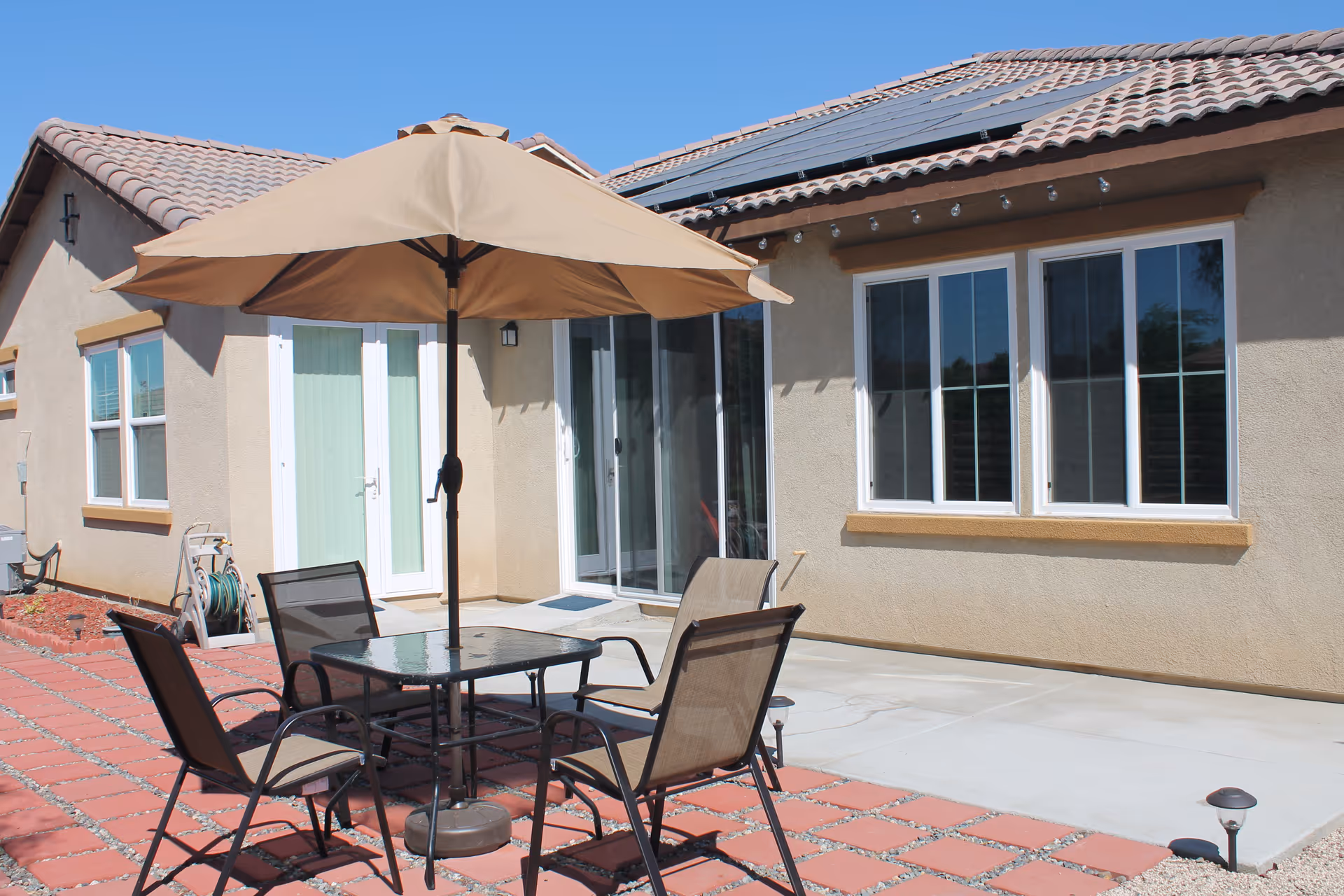 Outdoor patio area with a glass-top table, four chairs, and a large beige umbrella. The patio is adjacent to a beige stucco building with white-framed windows and sliding glass doors. The roof has solar panels and the ground is paved with red bricks and concrete.