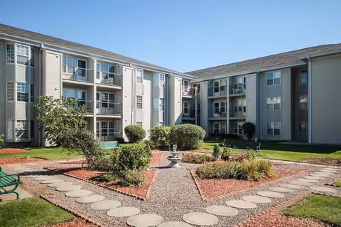 Landscaped courtyard with paved paths, benches and a small fountain surrounded by three-story apartment buildings with balconies under a clear blue sky.