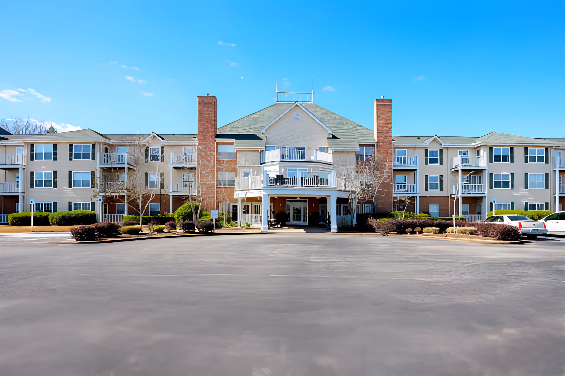 Three-story beige assisted living building with a central covered entrance, balconies and a wide paved parking area under a clear blue sky.