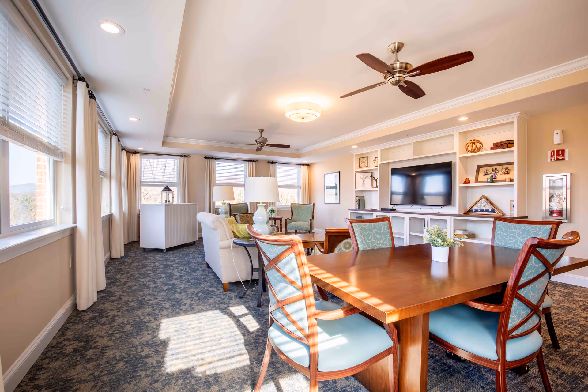 Bright common living area with a wooden dining table, teal chairs, and built-in shelves.