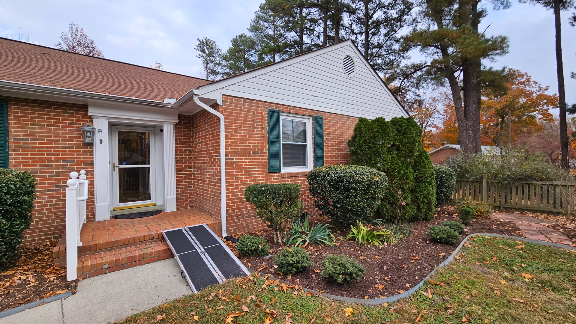 Brick senior living facility entrance with ramp, shrubs, and a wooden fence.