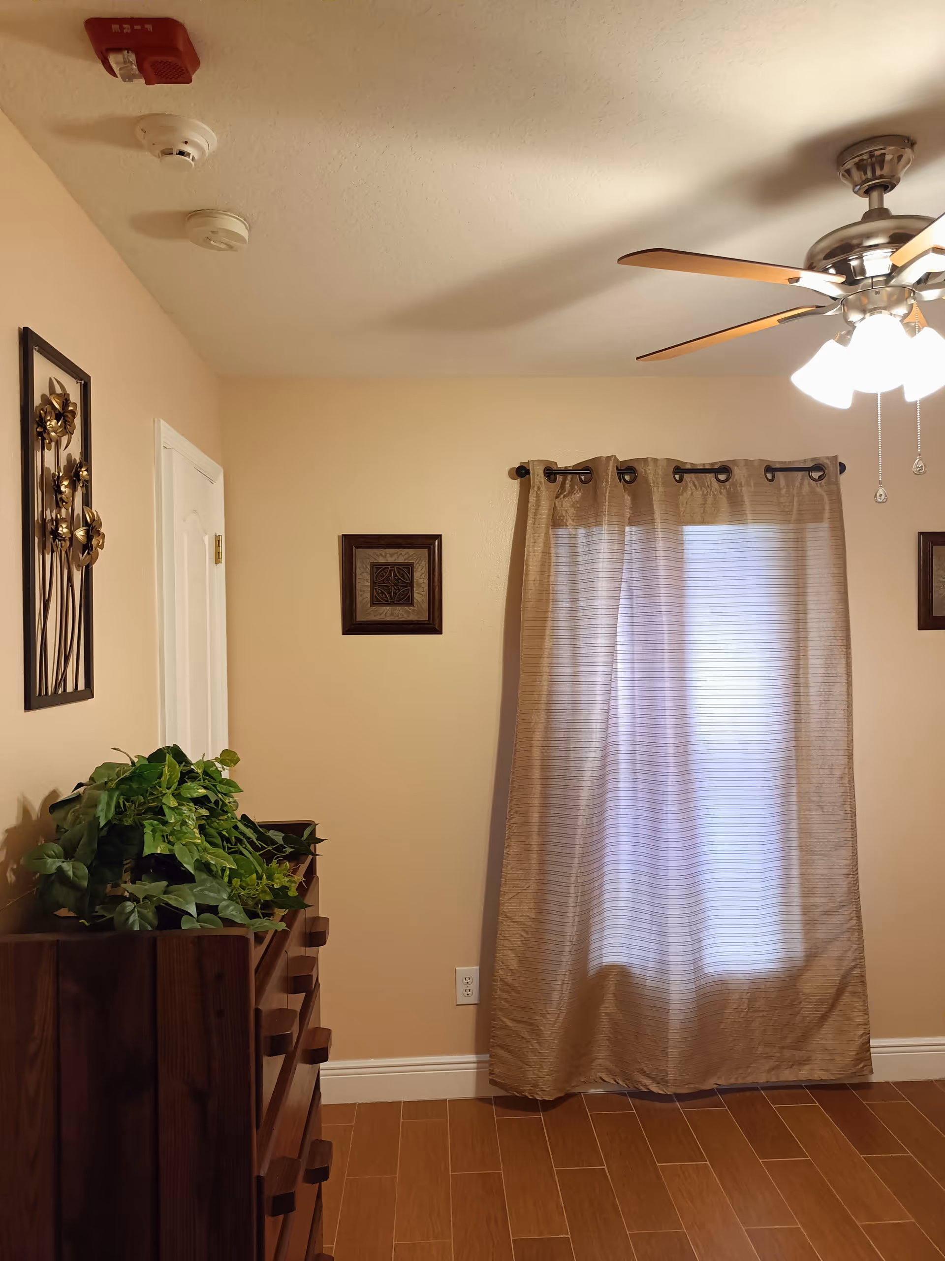 Corner of a beige living room with curtain, ceiling fan, and wooden dresser with a plant.