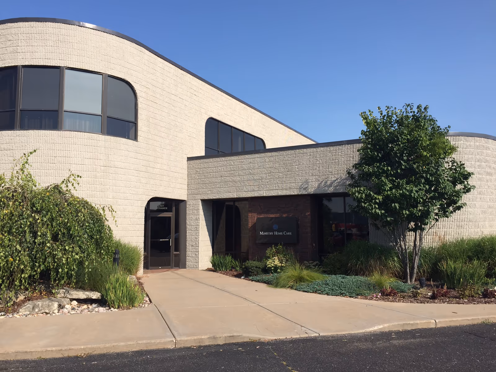 Curved beige building exterior with glass windows, entrance, and landscaped front.