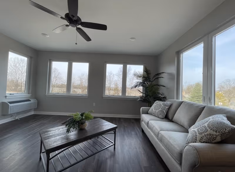 Bright common living room with sofa, coffee table, large windows, ceiling fan, and plants.
