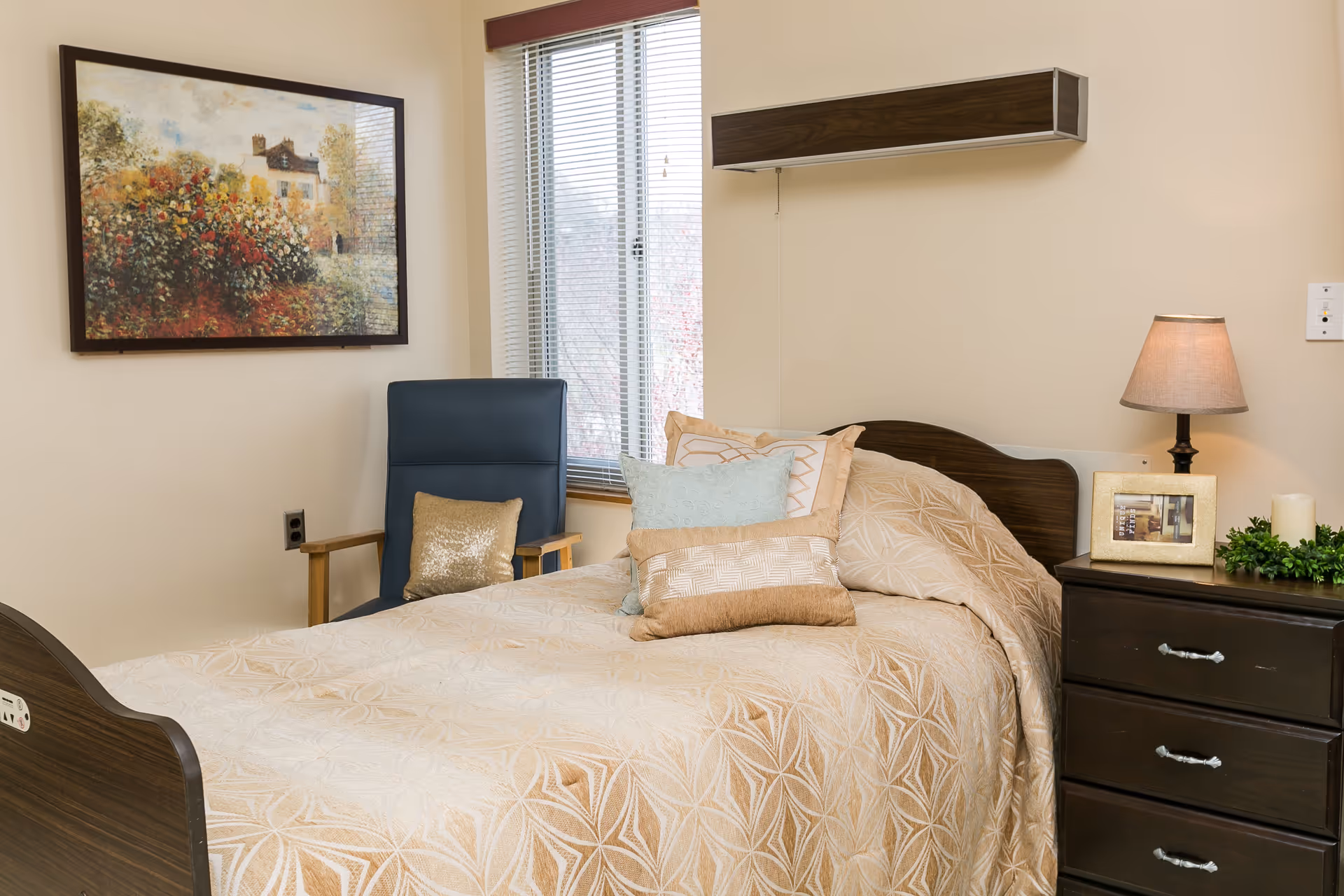 A furnished senior living bedroom with bed, nightstand, lamp, and window blinds.