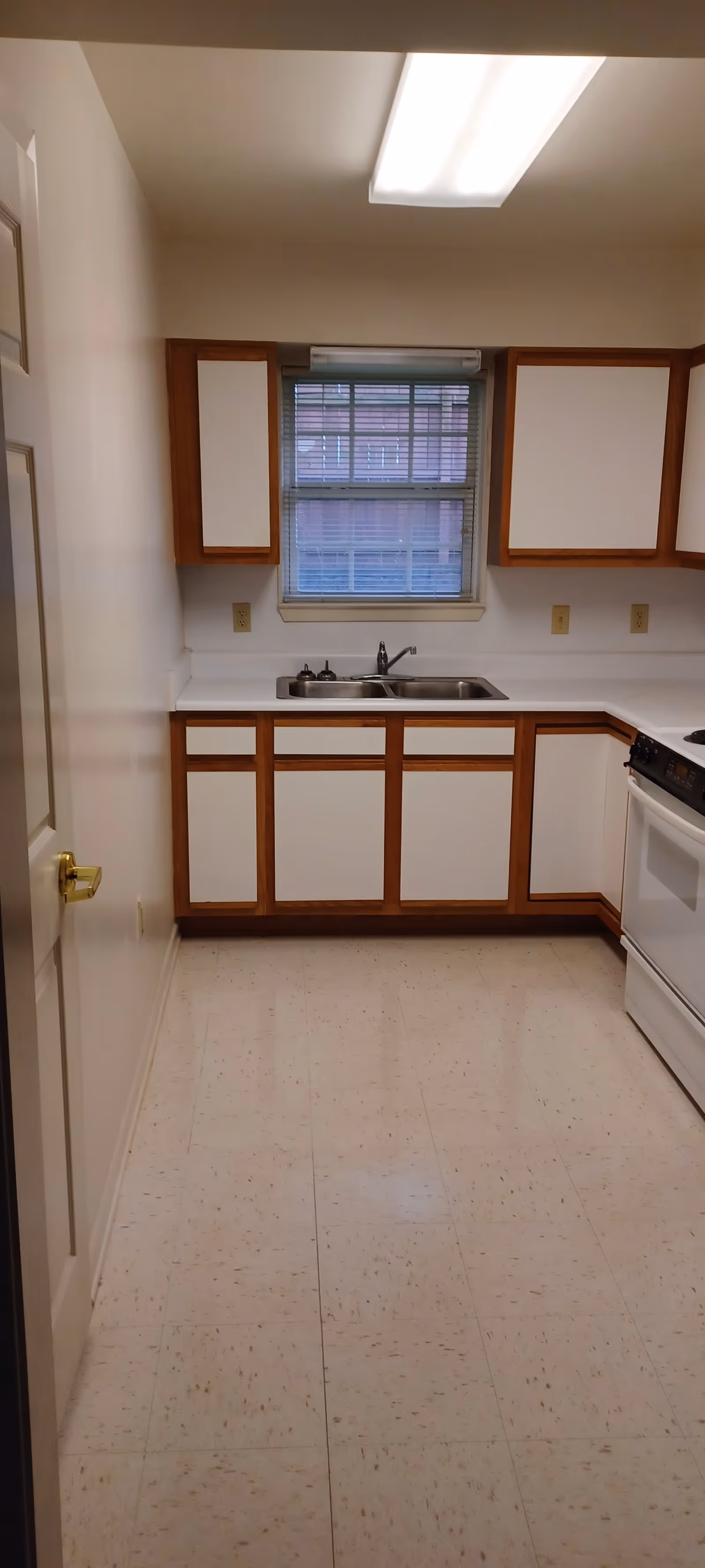 Narrow kitchen with white cabinets, brown trim, sink under window, stove on right.