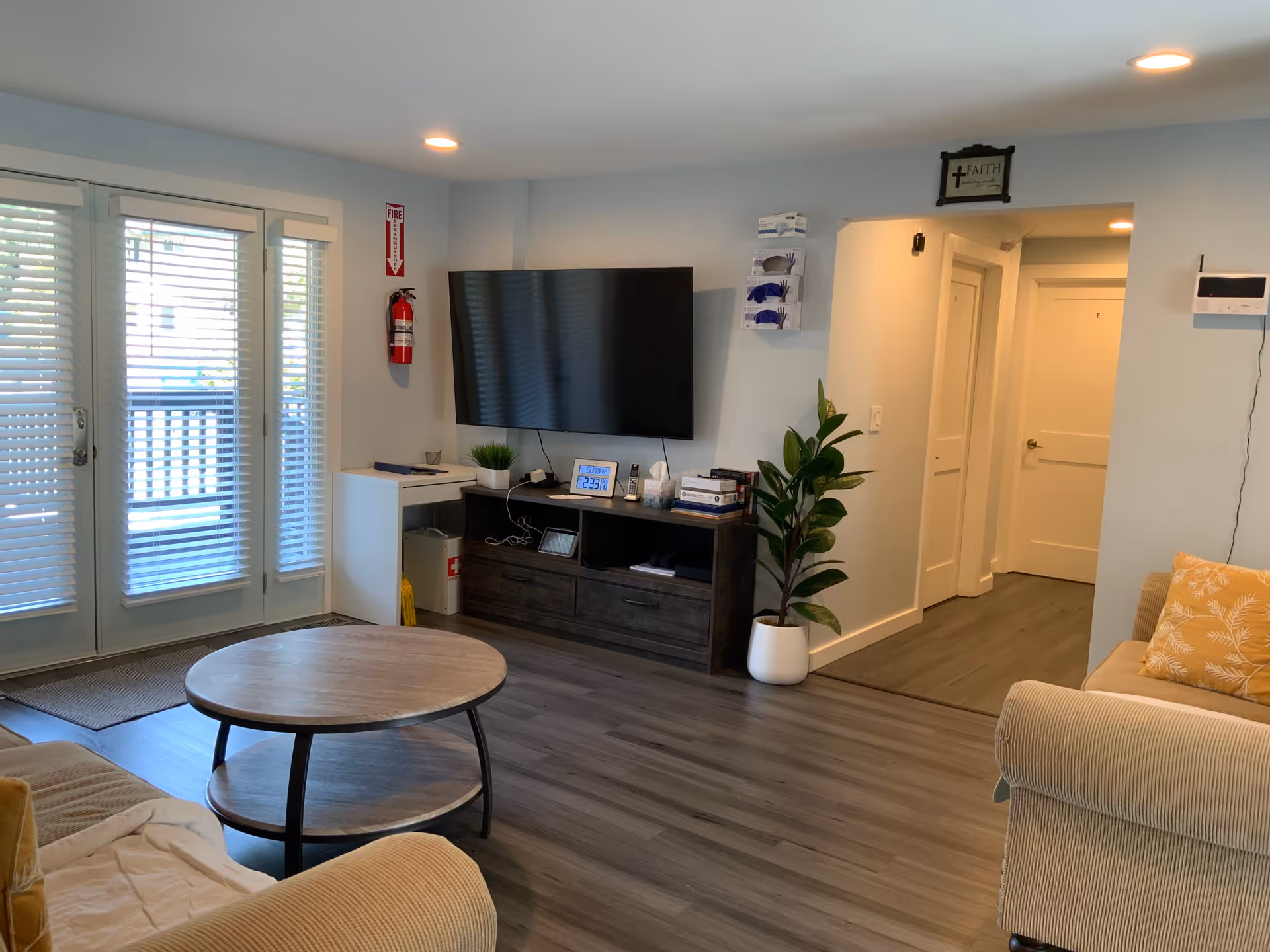 Bright living room with a wall-mounted TV, round coffee table, sofas, a potted plant and French doors with blinds.