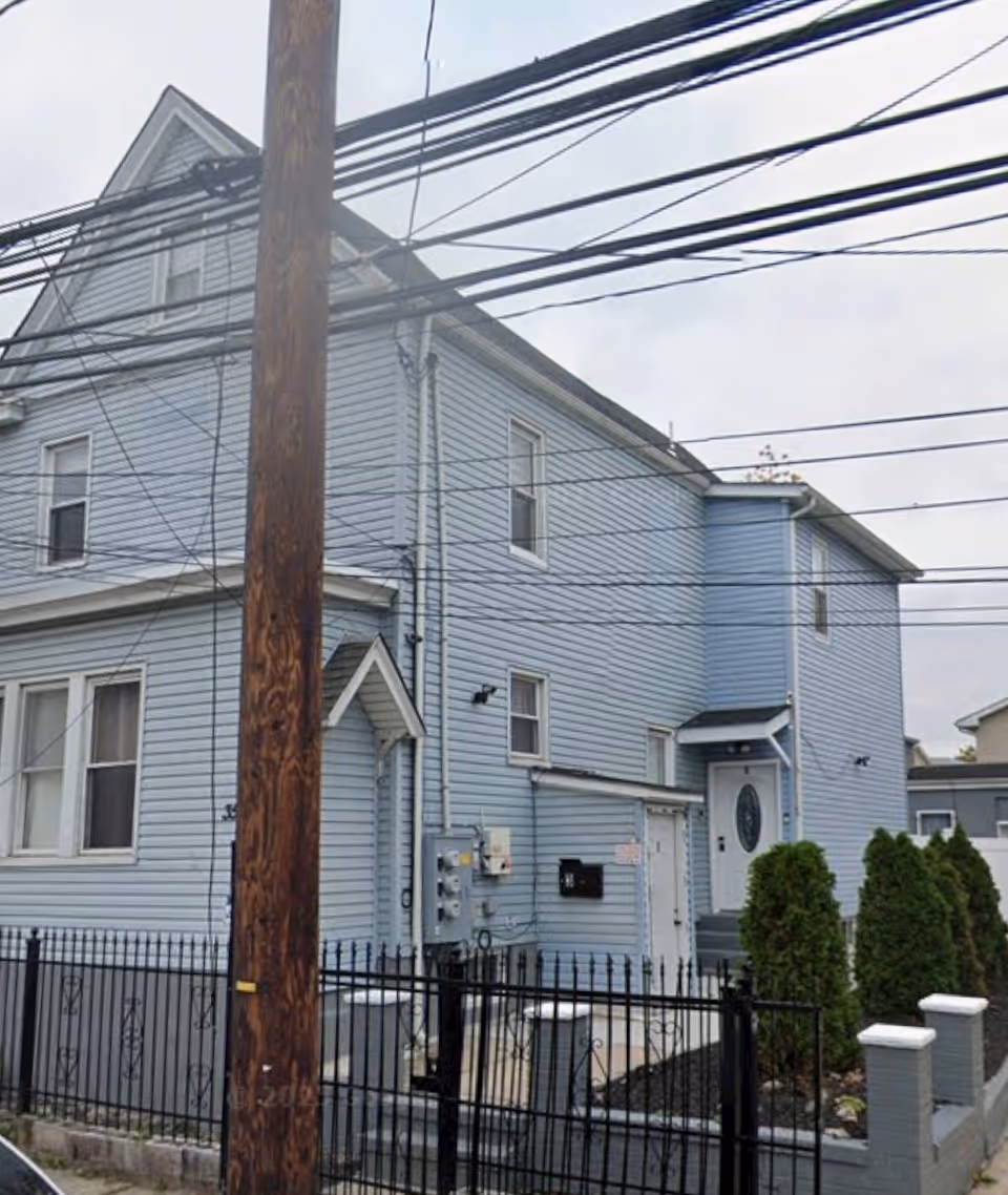 Two-story light blue building with front entry, gated yard, and overhead power lines.