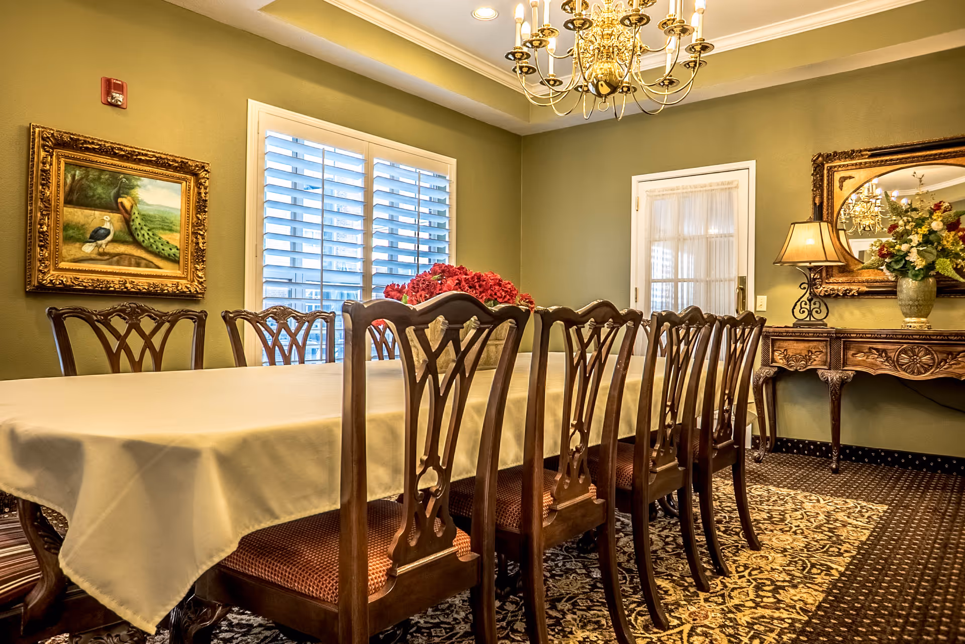 A formal dining room with a long table covered by a white tablecloth surrounded by wooden chairs with patterned cushions. The room features green walls, a large window with white shutters, a chandelier hanging from the ceiling, a framed painting of a peacock on one wall, and a decorative wooden side table with a lamp, a large mirror, and a floral arrangement.