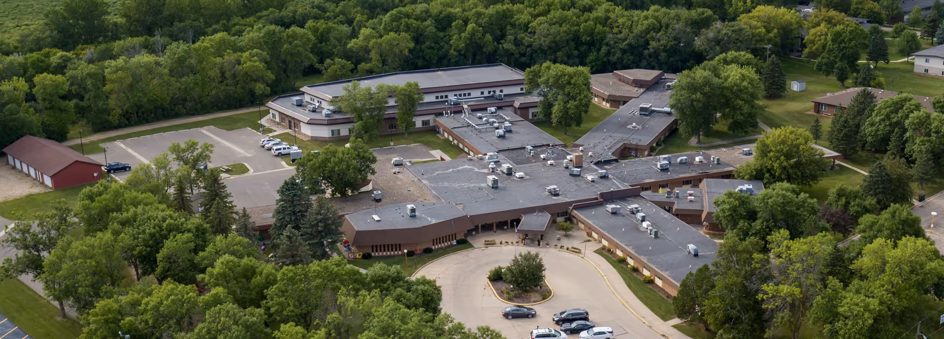 Aerial view of Morris Health Services DBA West Wind Village, showing a large, single-story building complex surrounded by trees and greenery. There is a circular driveway with parked cars in front of the building, and additional parking areas and smaller structures nearby.