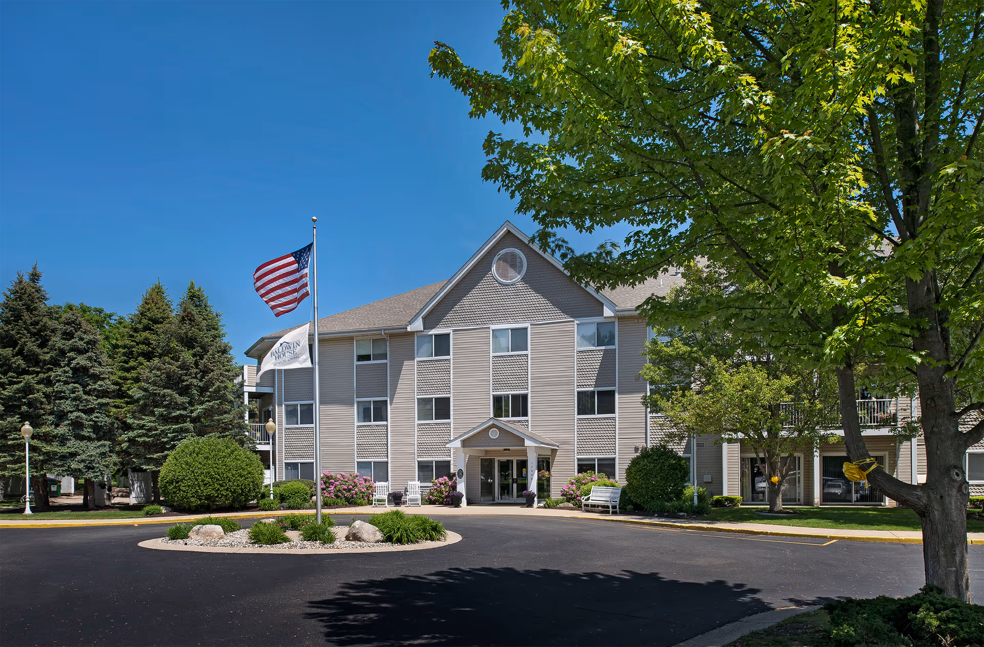 Exterior view of Baldwin House Senior Living Lloyd's Bayou building with trees, flags, and driveway.