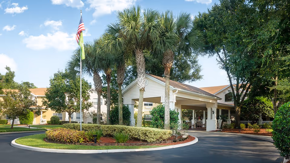 Exterior view of Ansley Parke at Orange City showing a driveway roundabout with palm trees and an American flag. The building has a covered entrance with white pillars and is surrounded by greenery and trees under a blue sky with some clouds.