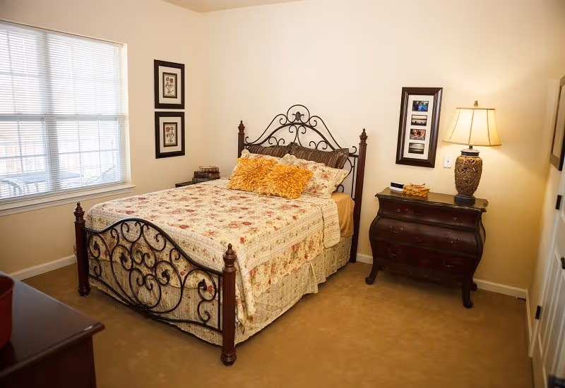 A bright bedroom with a decorative wrought-iron bed dressed in floral bedding, a nightstand with a lamp, and a window.