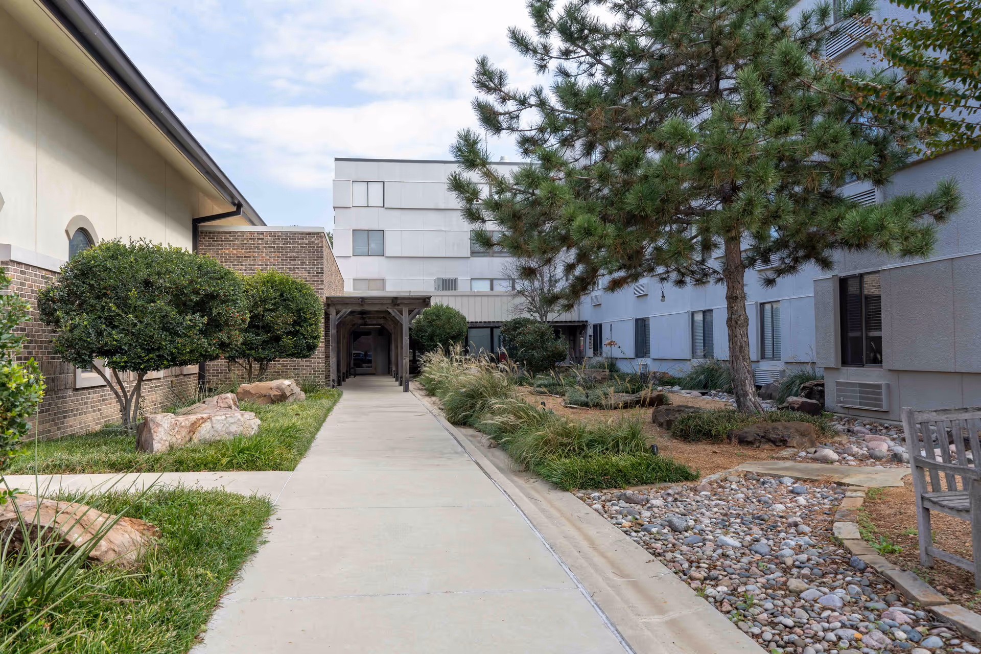 Exterior walkway of Morrison Residence featuring landscaped courtyard, trees, rocks, and a bench.