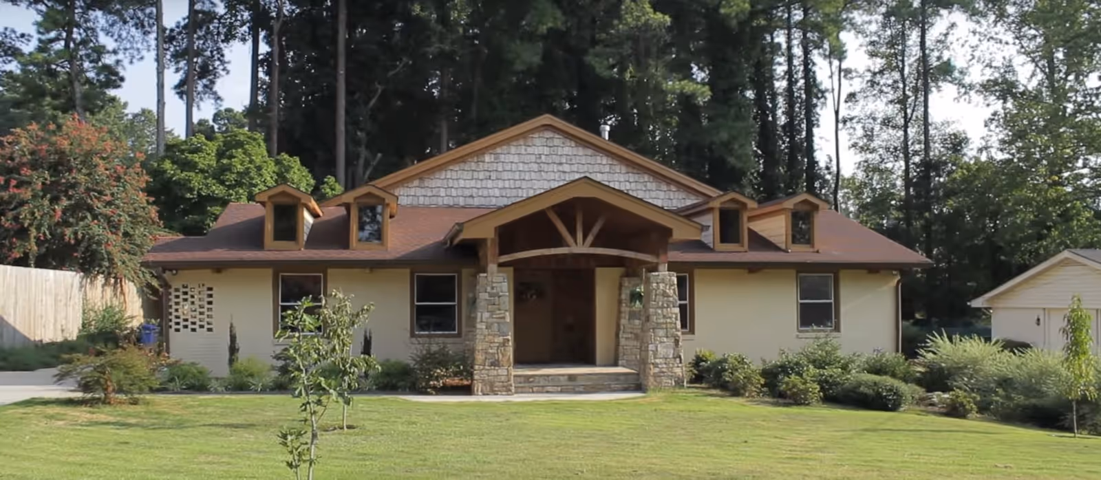 Single-story ranch-style building with a covered front porch supported by stone pillars, a manicured lawn and trees in the background.
