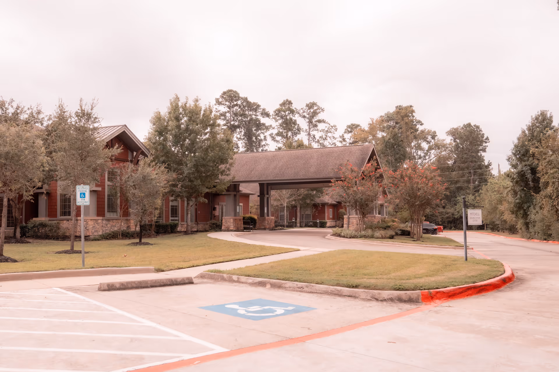 Exterior view of The Broadmoor At Creekside Park facility showing a covered entrance with a driveway, surrounded by trees and landscaping. There is a handicapped parking space in the foreground and a sign indicating reserved parking for accessible vehicles.