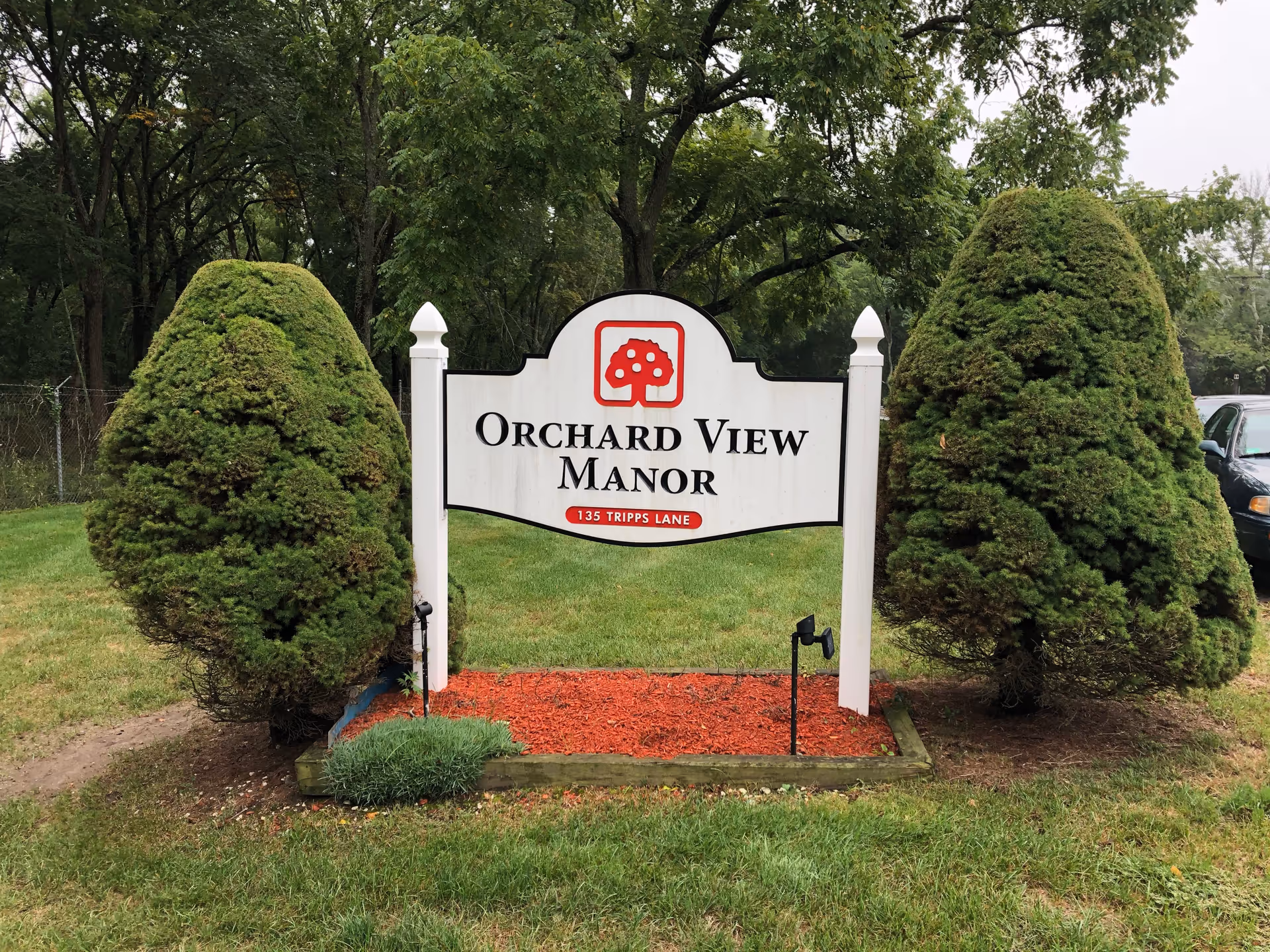 White wooden sign reading 'Orchard View Manor' with a red tree logo set between two trimmed evergreen shrubs on a lawn.