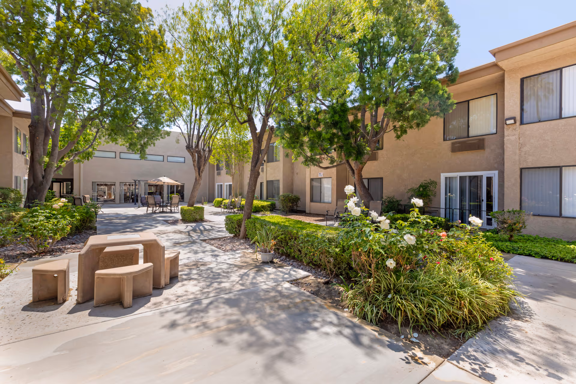 Outdoor courtyard area of a senior living facility with concrete pathways, green trees, bushes, and flowering plants. There are outdoor tables and chairs with umbrellas, and beige two-story buildings with windows surround the courtyard.