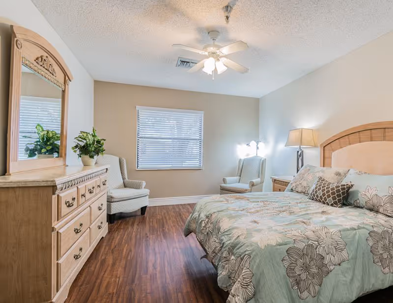 A furnished senior living bedroom with a bed, dresser, mirror, chairs, and ceiling fan.