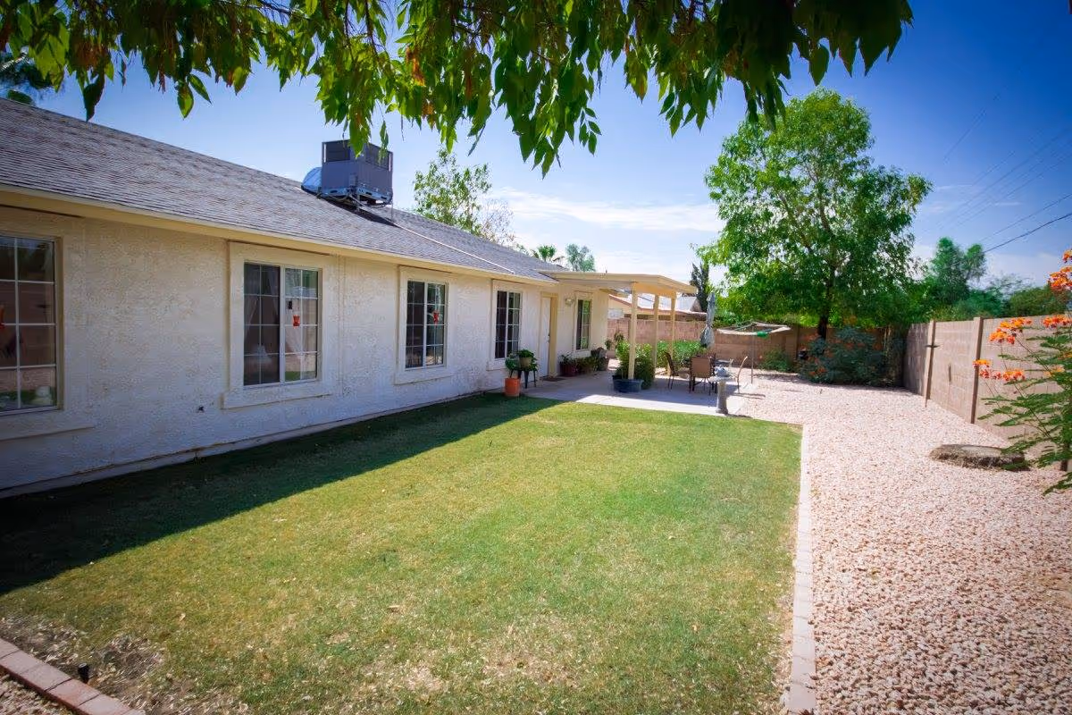 Outdoor view of a single-story assisted living facility with a grassy lawn, a gravel pathway, and a covered patio area with outdoor seating. Trees and plants surround the yard, and the sky is clear and blue.