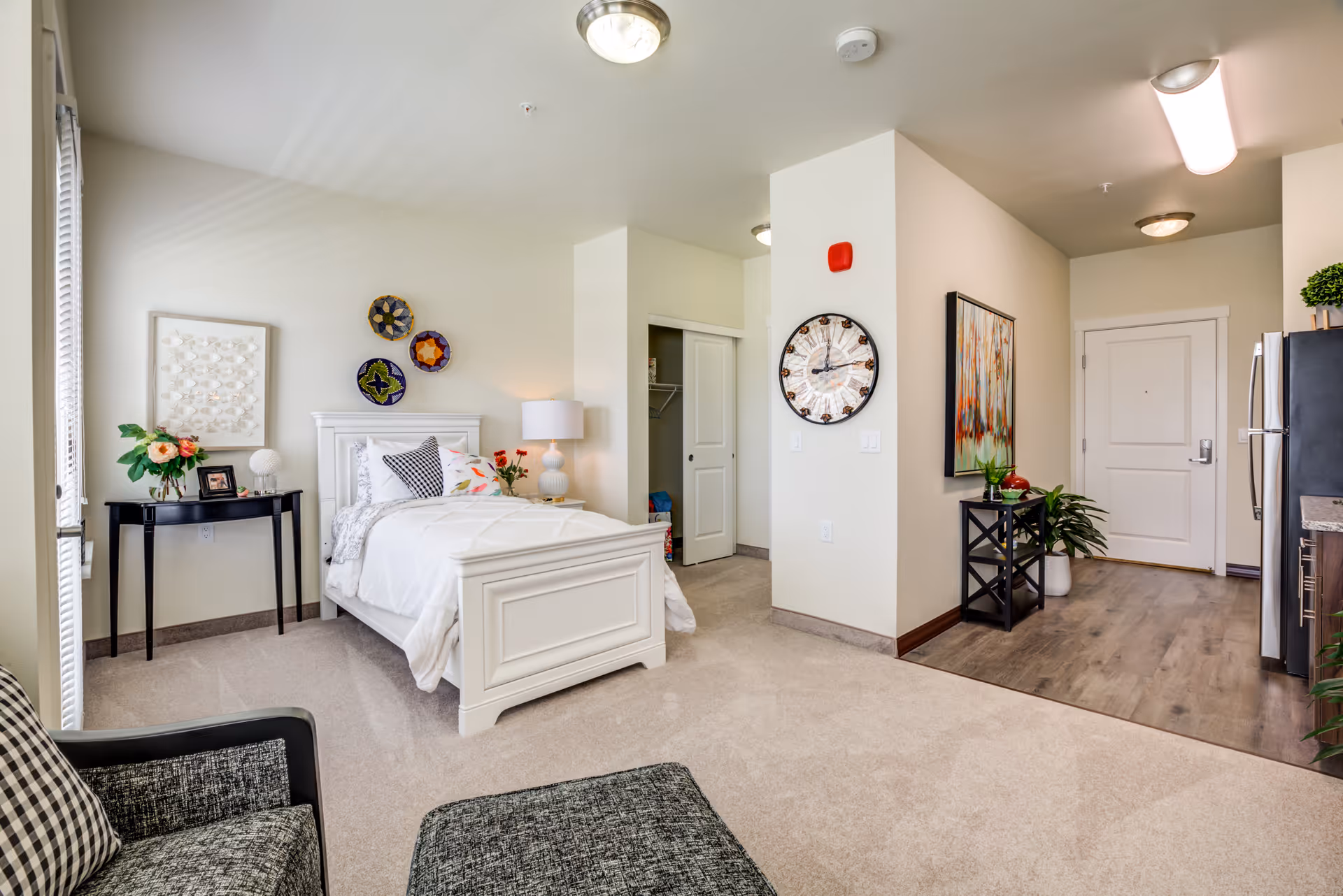 Bright studio apartment interior featuring a white bed, side table, seating area, and an entryway with a wall clock and artwork.
