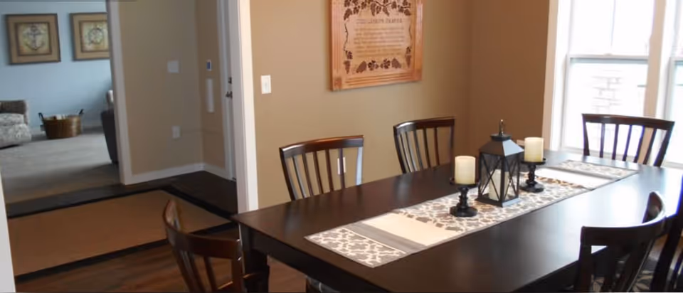 A dining room with a dark wooden table and six matching chairs. The table is decorated with a patterned runner, two candles on black holders, and a black lantern centerpiece. The room has beige walls, a framed decorative piece on the wall, and large windows letting in natural light. In the background, there is a view into a living area with carpeted flooring and furniture.
