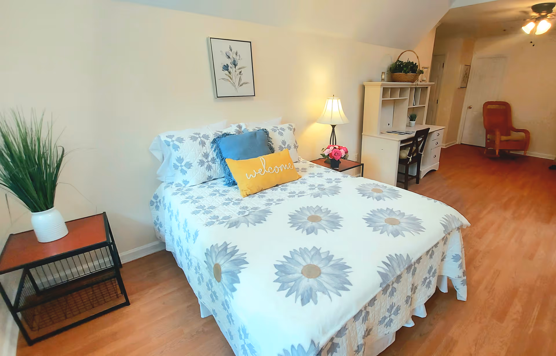 A cozy senior living bedroom with floral bedspread, desk, plants, and hardwood floor.