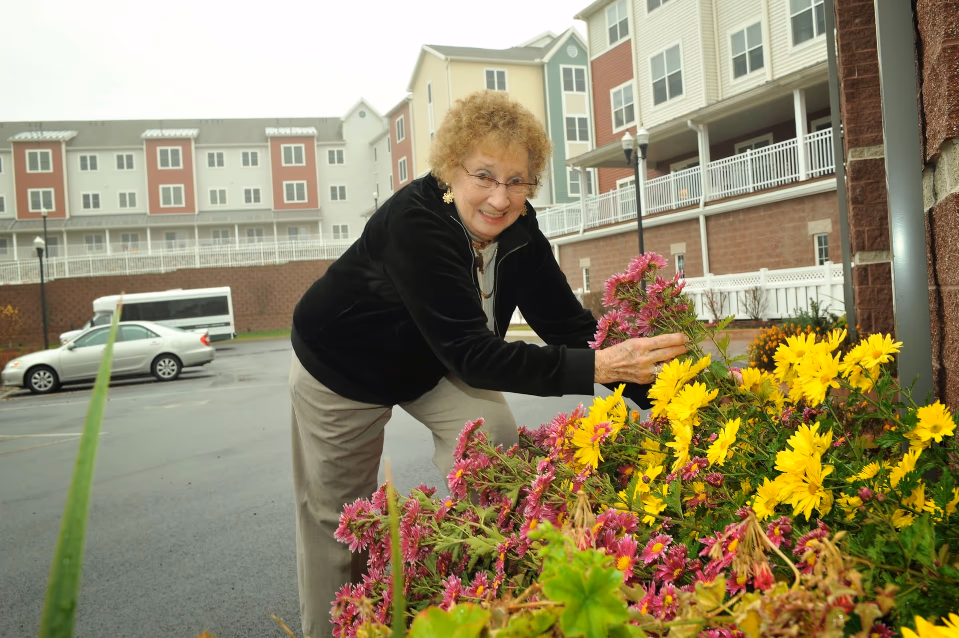 An elderly woman with curly hair and glasses is smiling while tending to a flower bed with yellow and purple flowers outside a multi-story residential building. There are parked cars and a white fence in the background.