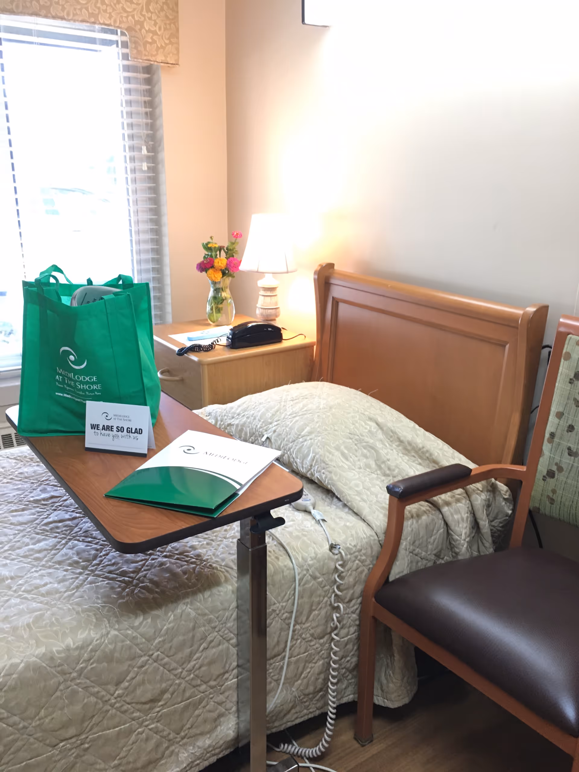 A furnished senior living bedroom with a bed, nightstand, lamp, chair, and green tote bag.