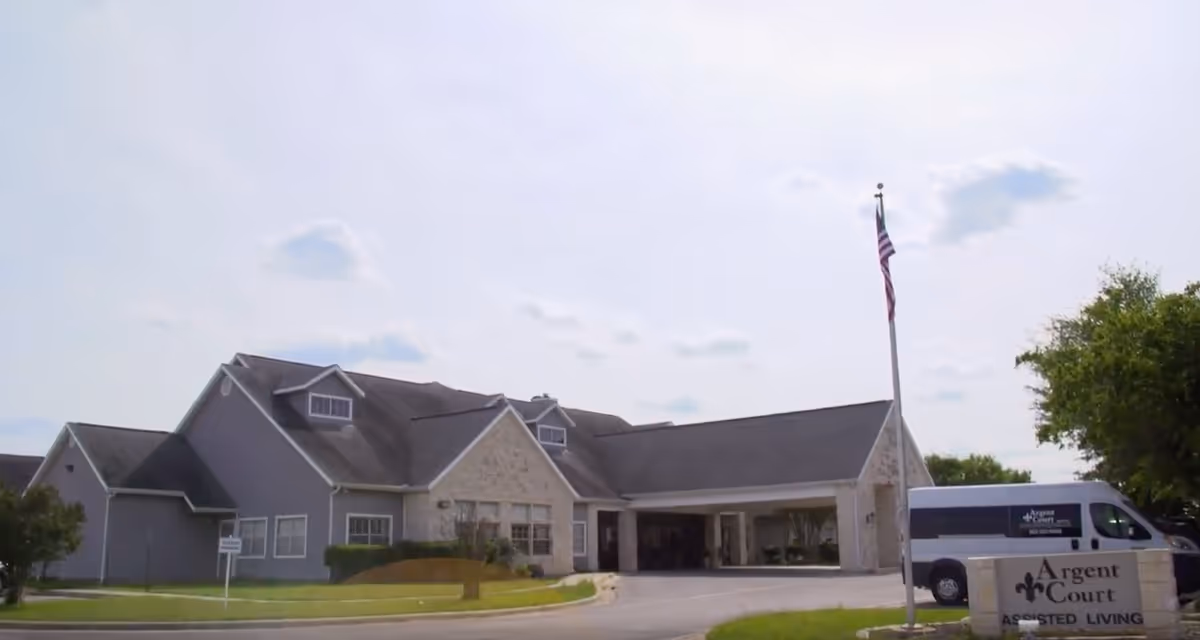 Front exterior of Argent Court Assisted Living showing the building entrance, an American flag, a van, and a stone sign.