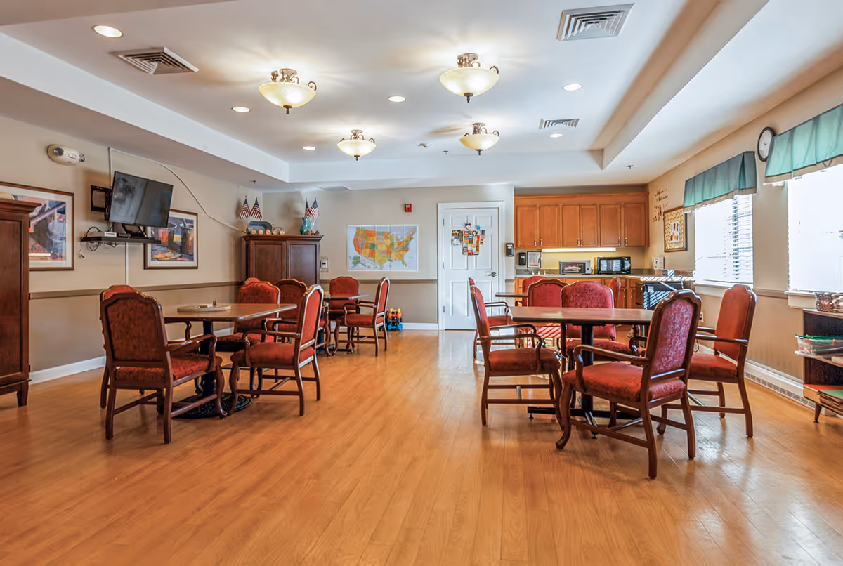 A bright senior living dining room with round tables and red upholstered chairs