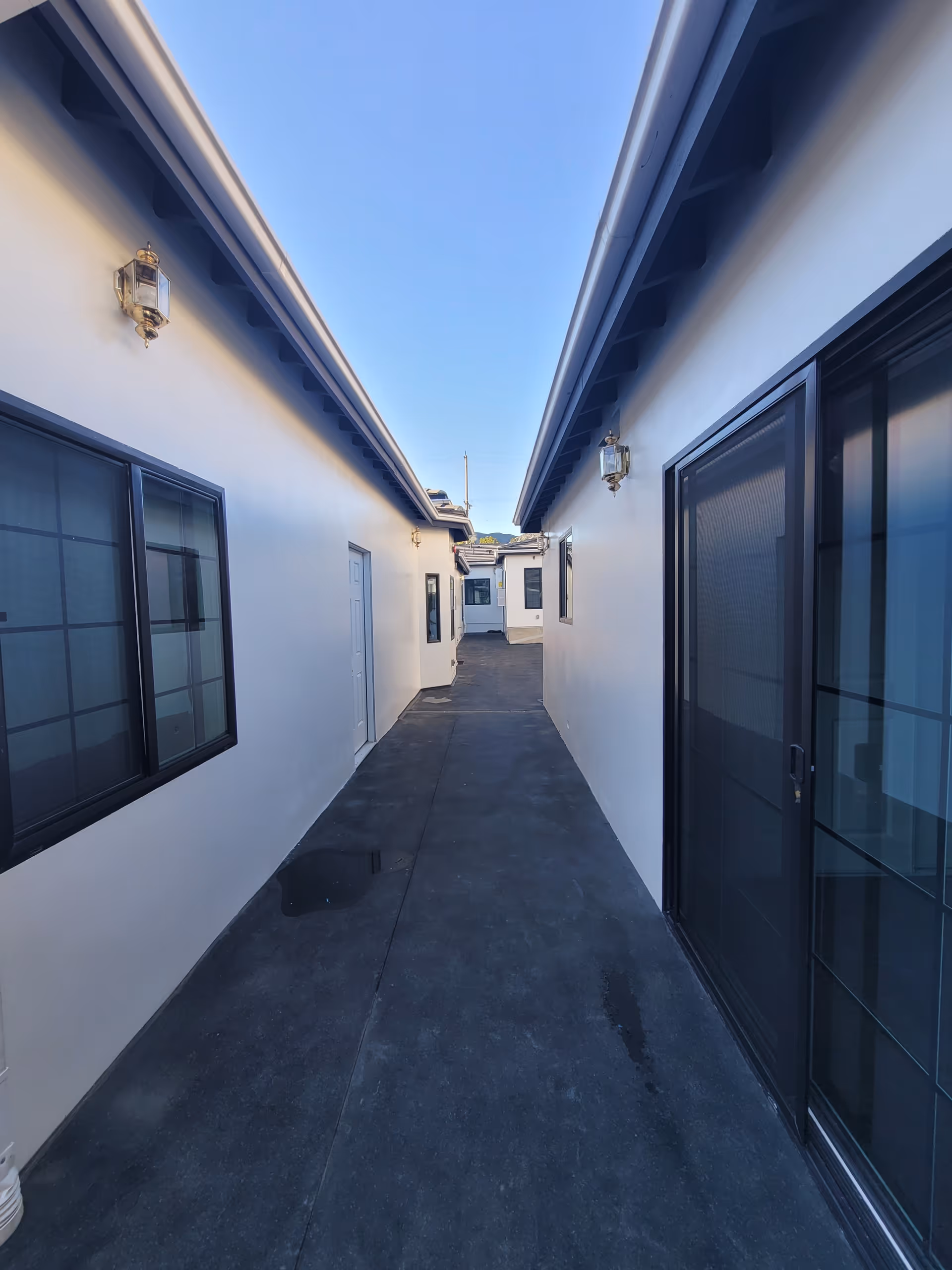 Narrow outdoor walkway between two white buildings with black-framed windows and doors, under a clear blue sky.